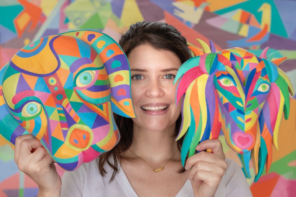 Artist holding two hand-painted geometric dog masks in front of a mural