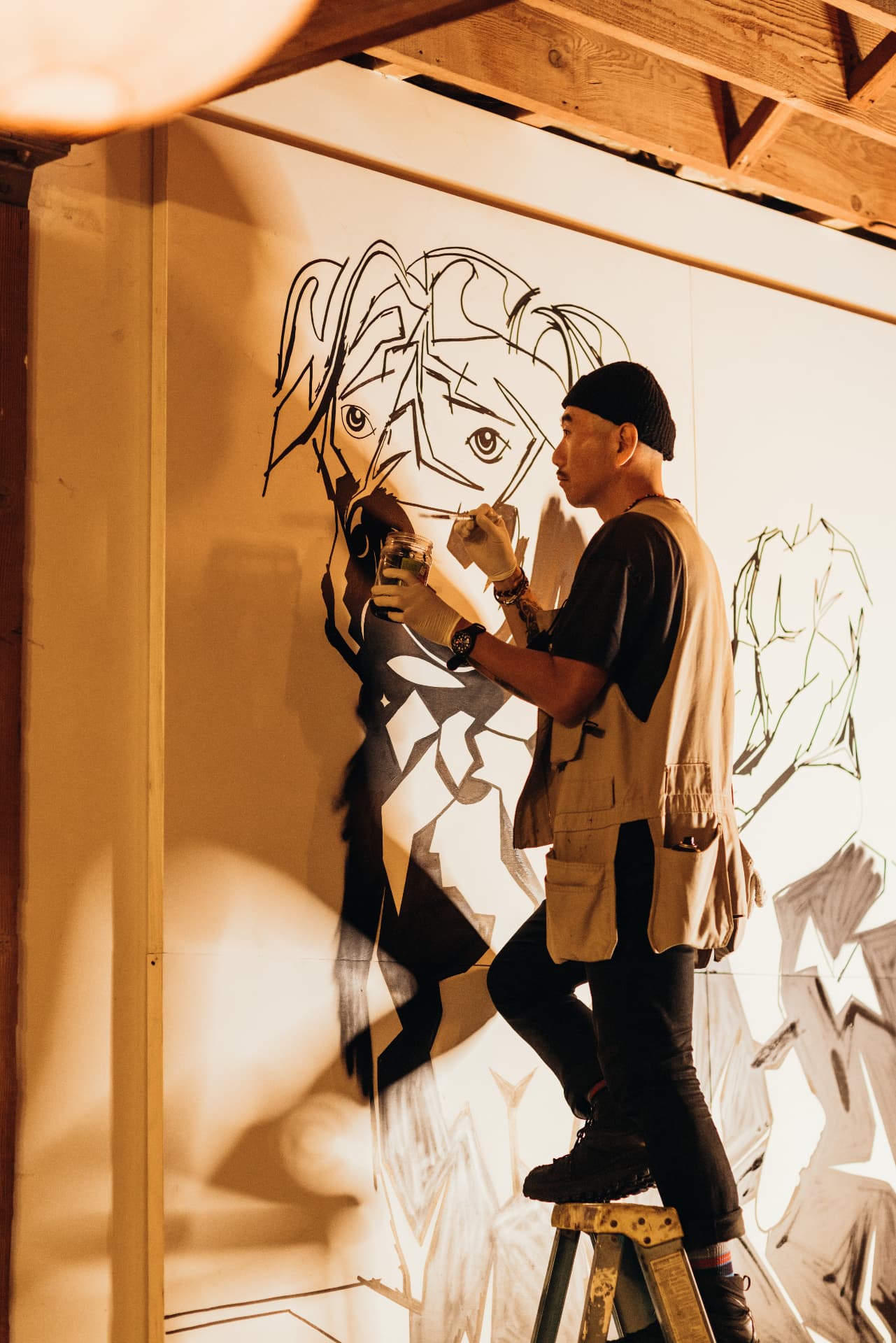 Artist on a ladder inking a large line-drawn mural under warm light